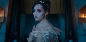 Mirren Mack as Empress Merwyn in The Witcher: Blood Origin. She wears a white gown made of butterfly wings. Her face is painted white, and the area around her left eye is painted magenta. She has gold leaves in her hair, which is tied up.