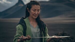 Michelle Yeoh as Scian in The Witcher: Blood Origin. She is standing in a desolate rocky landscape, wearing a gray-green coat over a gray tunic and holding a sword with both hands, smiling down at it.