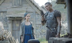 Bronwyn and Arondir from The Rings Of Power, standing at a forge while Arondir holds a black sword-hilt. He is wearing gray armor made from wood, with a leering face emblazoned on his breastplate. Bronwyn wears a simple blue dress and a heavy gray coat.