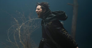 Ben Barnes as The Darkling in Shadow And Bone, standing in a wasteland with a dark sky, wearing a black hooded kefta with faint gold embroidery. His hood is thrown back, and he is screaming.