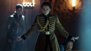 Jessie Mei Li as Alina Starkov in Shadow And Bone, standing in a dimly-lit tunnel with her arms outstretched to either side, staring down the camera. She wears a brown military uniform with gold stripes across the front and her dark hair is tied back.