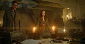 Archie Renaux as Mal and Jessie Mei Li as Alina in Shadow And Bone. They are standing in the dimly-lit cabin of a ship, but Alina illuminates the small room with balls of golden light hovering above her hands.