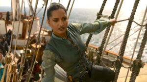 Amita Suman as Inej Ghafa in Shadow And Bone, holding onto the rigging of a ship with one arm. Her hair is tied back in a ponytail, and she wears a blue-green vest and leather pants.