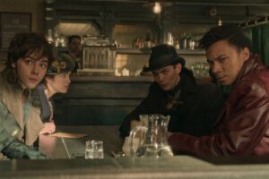 (left to right) Jack Wolfe as Wylan Van Eck, Danielle Galligan as Nina Zenik, Freddy Carter as Kaz Brekker, and Kit Young as Jesper Fahey in Shadow And Bone, seated around a table in a diner, all turning to look at the camera. Kaz wears a black bowler hat and overcoat, Jesper a maroon jacket, Nina a  dainty hat perched on the side of her head, and Wylan a waistcoat.