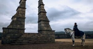 Moiraine Damodred, wearing a blue riding-cloak, sits on a white horse in a field of short grass looking up at two monumental brown stone pillars standing on a low plinth with steps carved in it. It appears to form a gateway, yet there is nothing on either side of it.