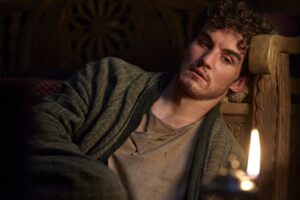 Mat Cauthon, lying on a wooden couch with his head leaning against the arm-rest, a single tear running down his left cheek. He is wrapped up in his thin, ragged green coat, and stares blankly at a candle-flame in the foreground.