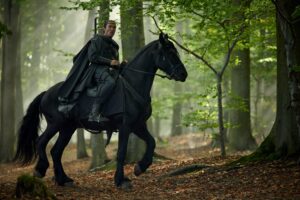 al'Lan Mandragoran, wearing a black cape over dark walking-clothes, riding a black horse through a forest. He has a sword strapped to his back, and an expression of surprise on his face.