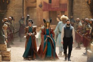 (left to right) two Seanchan women, Loial, and Ishamael, striding across a sandy courtyard in Falme between rows of Seanchan warriors and civilians. The two women wear long-sleeved rust-colored jackets and coats over teal blue gowns. The woman on the far left wears a sinuous metal face-mask. The woman in the center has her hair shaped into wings on either side of her head, and wears a golden mask covering her entire face except her chin. Ishamael wears a black vest over a white shirt and black trousers, while Loial, behind him, wears a light blue-green tunic.