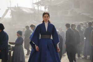 Moiraine Damodred in The Wheel Of Time season two, wearing a wide-sleeved dark blue dress over a white shirt, with a blue-and-gold belt. A gold circlet is nestled in her brown hair. She stands in a dusty marketplace, amidst a crowd of people.