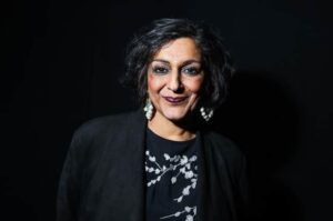 British actress and comedian Meera Syal, standing against a dark background. She has short dark messy hair, dark eyeshadow and lipstick, and smiles as if she knows a secret. She is wearing a black blazer.