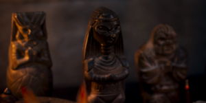 Three votive statues displayed prominently in The Wheel Of Time's first season, carved from brown stone, standing on a mantel-piece. The statue on the far left depicts a woman with a square headdress, whose dress has a web-like pattern. The middlemost statue depicts a woman with long hair and a collar of rings. The statue on the far right depicts a short, stout man with a beard and an angry expression.