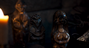 Three votive statues, prominently displayed in The Wheel Of Time's first season, carved from brown stone, standing on a mantel-piece surrounded by burning candles. The statue on the far left depicts a tall man holding a guitar. The middlemost statue depicts a short man with vague, angular features. The statue on the far right depicts a woman with long hair, a large bosom, and wide hips.