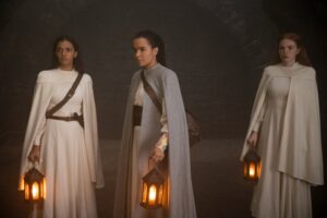 (left to right) Madeleine Madden as Egwene, Zoe Robins as Nynaeve, and Ceara Coveney as Elayne in The Wheel Of Time, wearing nearly identical white gowns with capes, standing in a dark cellar holding lanterns. Nynaeve's dress has stripes of vibrant color on the sleeves, and she wears a prominent gold ring on her left hand.