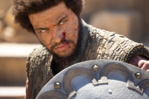 Marcus Rutherford as Perrin Aybara in The Wheel Of Time, bloody and bruised, glowering with bright golden eyes at something offscreen. He wears lightweight leather armor over a pale red shirt, and is carrying a round studded shield in front of him.