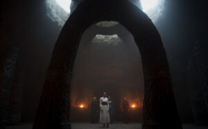 Nynaeve al'Meara, wearing a white dress with a wide leather belt, standing framed between the stone pillars of a silver archway standing on a dais in the center of a round stone chamber underneath the White Tower. Candles burn in sconces on the far wall. Behind Nynaeve are Sheriam Bayanar, Leane Sharif, and Liandrin Guirale.