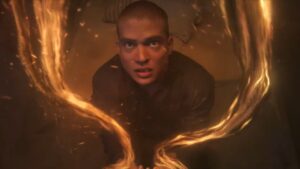 Josha Stradowski as Rand al'Thor in The Wheel Of Time, wearing dark clothes, kneeling on the floor of a small bedroom in an inn, staring up with wide, horror-stricken eyes as orange threads of the One Power burst from his hands and curl upwards around him towards the ceiling.