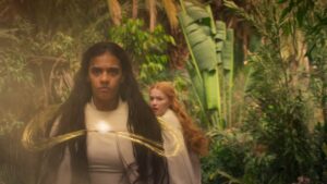 Madeleine Madden as Egwene al'Vere in The Wheel Of Time season two, standing in a tropical forest with Ceara Coveney as Elayne Trakand just behind her. Both wear plain white dresses and cloaks. Egwene's dark hair is loose, and she is facing down an unseen opponent, channeling golden threads of the One Power to form a levitating hoop encompassing her, while Elayne is turning to run away, with a frightened expression on her face.