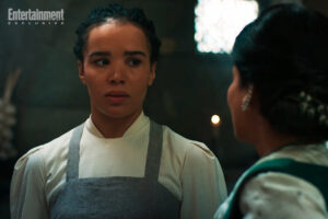 Zoe Robins as Nynaeve al'Meara in The Wheel Of Time season two, standing in the White Tower kitchens wearing a plain gray apron over a long-sleeved white dress, with her hair pulled into a braid. She has a troubled expression on her face. In the foreground, Priyanka Bose as Alanna Mosvani stands facing her, away from the camera, wearing a dark green vest over a white blouse, with her dark hair tied up in a bun adorned with green ornaments.