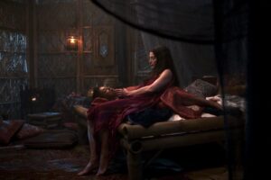 Rosamund Pike as Moiraine and Sophie Okonedo as Siuan in The Wheel Of Time, lying together in a wooden bed wearing red nightgowns. Moiraine is sitting up slightly. while Siuan is gazing up at her. They are in a low wooden hut with fishing-nets hanging from the ceiling.