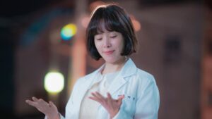 Han Ji-min in Behind Your Touch, wearing a white veterinarian's coat and looking down at her hands with a slight smile on her face.