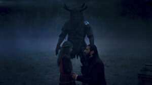 Ishamael in The Wheel Of Time season two, played by Fares Fares, wearing a black coat with white cuffs, kneels down to be on eye-level with a small girl in a red-and-blue dress. Standing between them, towering over both, is a Trolloc wearing bits and pieces of leather armor, covered in fur, with broken antlers and tusks protruding from its face. It is nighttime, and fog is drifting over the short grass through a courtyard filled with stone pedestals.