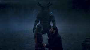 Fares Fares as Ishamael in The Wheel Of Time season two, wearing a dark coat, kneeling in short grass while holding the hands of a small girl wearing a red and blue dress. Behind them looms a massive Trolloc, man-shaped but with the face of a boar, with antlers sprouting from its head. It is night, and fog is rolling in.