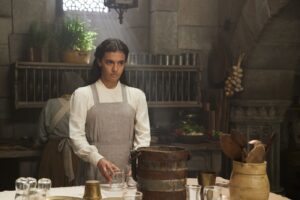 Madeleine Madden as Egwene al'Vere in The Wheel Of Time season two, standing before a table in a dimly-lit kitchen, wearing a gray apron over a long-sleeved white gown.