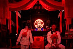 Greg Hsu in Marry My Dead Body, wearing a white suit, sitting alongside a similarly-dressed mannequin beneath a red canopy in a darkly-lit room.