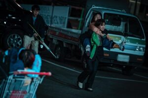 Lee Si-Young carrying Park Na-rae on her shoulders in Zombieverse as they run through a parking lot at night pursued by zombies.