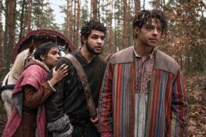 (left to right) Madeleine Madden as Egwene al'Vere, Marcus Rutherford as Perrin Aybara, and Daryl McCormack as Aram from The Wheel Of Time season one, standing in a forest. Egwene is wearing a purple-pink shawl over a red-brown coat, looking anxiously over Perrin's shoulder. Perrin is wearing a dark brown fur coat with a leather  satchel. Aram is wearing a striped jacket in muted shades of red, blue and yellow over a white shirt with floral embroidery.