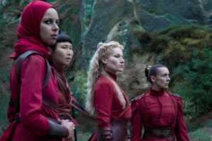 Kate Fleetwood as Liandrin Guirale in The Wheel Of Time season one, wearing red and brown, with her long white-blond hair flowing down her back in a mane of intricate braids, standing surrounded by similarly-dressed women in a rugged landscape.