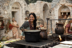 Priyanka Bose as Alanna Mosvani in The Wheel Of Time, standing in her outdoor kitchen and smiling as she leans over a large pot.