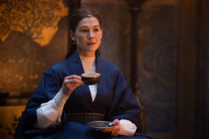 Rosamund Pike as Moiraine Damodred in The Wheel Of Time, sitting in a room with gold-and-black paneled walls. She has a saucer resting on her lap, and is lifting a black tea-cup to her lips. She is wearing a wide-sleeved dark blue robe cinched at the waist with a wide blue-and-gold cloth belt over a long-sleeved white knitted blouse. A single blue gemstone sits on a slender golden diadem above her brow. The Great Serpent ring on her left hand sports a larger, darker, polished blue gemstone.