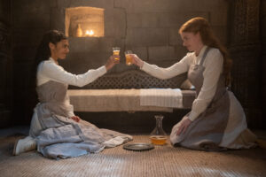 Madeleine Madden as Egwene al'Vere sitting across from Ceara Coveney as Elayne Trakand in a small room with stone walls, lit by candles burning in niches. Both are wearing gray aprons over long-sleeved white gowns, and wear white boots. They are raising glasses filled with a light-orange beverage, and smiling.