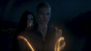 Josha Stradowski as Rand al'Thor and Natasha O'Keefe as Lanfear in The Wheel Of Time, standing outside at night. Rand is channeling golden threads of the One Power that wind around his arms, while Lanfear hovers over his right shoulder, her face partially obscured by her long jet-black hair.