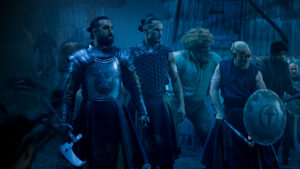 (left to right) Gregg Chilingirian as Ingtar, Arnas Fedaravicius as Masema, Hammed Animashaun as Loial, and Guy Roberts as Uno from The Wheel Of Time, standing with weapons drawn in a village at night. Ingtar, Masema, and Uno wear light armor.