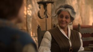 Meera Syal as Verin Mathwin in The Wheel Of Time, sitting in a chair outdoors with a smile on her face. She has gray hair bunched up on top of her head, with loose curly strands falling around her face. She is wearing a silk brown dress clasped at the front with a large golden teardrop-shaped brooch, over a white blouse with long, frilly  pleated sleeves.