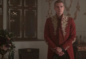 Josha Stradowski as Rand al'Thor in The Wheel Of Time, standing with his hands folded in a room with fancily decorated walls. He is wearing a high-collared, long-sleeved dark red coat with golden herons in flight embroidered around the cuffs and collar, over a red vest and dark brown trousers. His head is shaven.