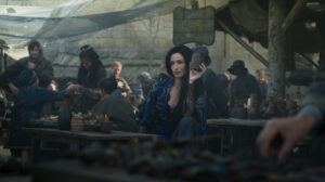 Natasha O'Keefe as Selene in The Wheel Of Time, sitting outdoors at a table underneath a wooden sign for her inn with a painted crescent moon. She has long black hair, and is wearing a dark blue robe over a lacy black dress, holding up a cup.