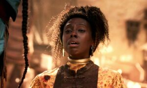 Nyokabi Gethaiga as Ryma in The Wheel Of Time, from the shoulders up. She is wearing a brown vest over a bloodstained yellow floral-patterned blouse, and has a gold collar around her neck. She has dark hair piled up on top of her head. She is sobbing.