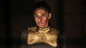 Madeleine Madden as Egwene al'Vere in The Wheel Of Time, standing in a dark room wearing a large golden collar and breastplate, all of one piece, over a long-sleeved gray dress. Her dark hair is pulled back into a braid. Her face is bloodied and bruised.