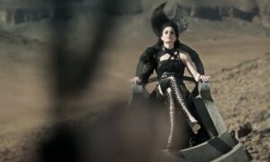 Natasha O'Keeffe as Lanfear in The Wheel Of Time, sitting with her legs crossed on a black stone throne in the middle of the desert. She is wearing a black dress that leaves her left arm bare, thigh-high black lace-up boots, and a black leather harness. A black lace crescent-moon headdress sits on her head. She has short jet-black hair.