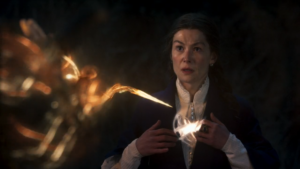 Rosamund Pike as Moiraine Damodred in The Wheel Of Time, wearing a dark blue gown over a high-collared knitted white blouse. She has her hands over her chest, where a glowing knot  of white light is hovering. From the foreground, a tentacle of black and orange light with a barb at the end is approaching her. She has a nervous expression on her face.