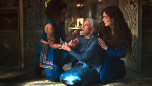 Sophie Okonedo as Siuan Sanche and Rosamund Pike as Moiraine Damodred, both wearing royal blue, kneeling on the floor supporting Hayley Mills as Gitara Moroso, an old woman in a blue dress with long white hair and white eyes, who has collapsed.