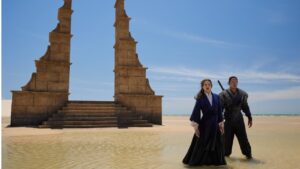 Rosamund Pike as Moiraine Damodred and Daniel Henney as Lan Mandragoran in The Wheel Of Time, standing in shallow water on a beach with clear skies above them. Behind them looms a sandstone gate on a dais with steps leading up to it. Moiraine is wearing a dark blue long-sleeved gown over a knitted white blouse. She has long brown hair. Lan is wearing a gray tunic and trousers, with a sword strapped to his back. His dark hair is worn in a topknot.