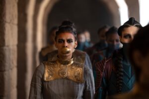 Madeleine Madden as Egwene al'Vere walking alongside Xelia Mendes-Jones as Renna through a stone colonnade. Egwene is wearing a large gold collar over a long-sleeved gray dress, and has a golden disc in her mouth as a gag. Her dark hair is braided. Renna wears lightweight red-brown leather armor like the carapace of a beetle over a dark blue long-sleeved shirt. Her dark hair hangs in two long braids, and the lower face of her face is painted dark blue.