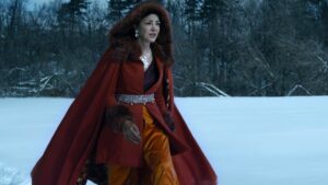 Shohreh Aghdashloo as Avasarala in The Expanse, striding through a snow-covered field wearing a fur-lined crimson cape with a hood over a jacket of the same color and mustard-yellow trousers. She has fur gloves, large earrings, and a silver belt.