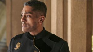 Alexander Karim as Lews Therin Telamon in The Wheel Of Time, standing in a sunny room with brown stone walls, staring off into the distance. He is wearing a black military-style uniform with gold embroidery, and a large golden dragon emblazoned on the right breast.