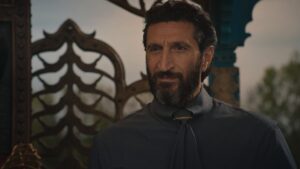 Fares Fares as Ishamael in The Wheel Of Time, wearing a gray handsomely tailored dress-shirt, with his dark hair slicked back. He is smiling slightly. He is standing on a palanquin, with a metal railing behind him.