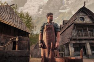 Marcus Rutherford as Perrin Aybara in The Wheel Of Time season one, standing in the Two Rivers with wooden houses behind him and a mountain vista. He is wearing a leather apron over a green-blue shirt with the sleeves rolled up. He has short dark curly hair.